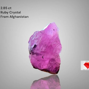 Könnte beinhalten: Ein roher, rosa Rubinkristall aus Afghanistan. Der Edelstein, mit einem Gewicht von 2,85 ct, hat eine unregelmäßige Form mit einer reflektierenden Oberfläche. Das Bild enthält Text, der die Herkunft und das Gewicht des Kristalls angibt.