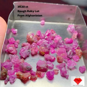 May include: A collection of rough ruby gemstones in a small metal tray. The rubies are various shades of pink and red, with a total weight of 39.20 carats. Text on the image reads "Rough Ruby Lot From Afghanistan."