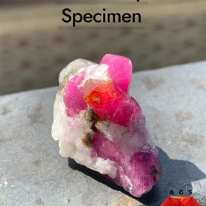 Pode incluir: Um espécime de rubi de 85 ct com estruturas cristalinas rosa vibrante e vermelhas incrustadas numa matriz mineral branca e cinzenta. O texto 85 ct Ruby Specimen é visível no topo, com um logótipo AGS e Afghan Gemstone Source.