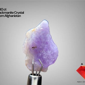 Puede incluir: Un cristal de Hackmanita de color púrpura claro de Afganistán, con un peso de 5,40 ct, es sostenido por una herramienta de metal. El cristal tiene una forma rugosa e irregular con una mezcla de tonos púrpuras y blancos. El texto dice "5.40 ct Hackmanite Crystal From Afghanistan."