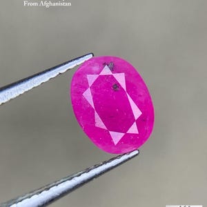 Puede incluir: Un rubí facetado de forma ovalada de Afganistán, sujeto con pinzas. La piedra preciosa es de color rosa vibrante con un patrón de facetas en forma de estrella. El texto dice "1,21 ct Rubí Facetado De Afganistán."