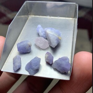 Op de afbeelding: Een verzameling van zeven ruwe, lichtpaarse en bleekroze hackmaniet edelstenen in een kleine metalen schep. De tekst 24.70 ct Hackmanite Rough Lot From Afghanistan staat bovenaan, met daaronder het logo van AGS Afghan Gemstone Source.