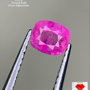 Puede incluir: Un rubí facetado de color rosa brillante, sujeto con pinzas. La gema tiene un patrón en forma de estrella en el interior. El texto en la parte superior dice "0.31 ct Faceted Ruby From Afghanistan". La imagen incluye un logotipo de Afghan Gemstone Source.