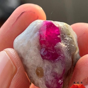 Puede incluir: Un espécimen de rubí de 54 ct que presenta un centro cristalino de color rojo rosado intenso y vibrante incrustado en una matriz mineral rugosa, opaca, de color blanco roto y gris. El texto 54 ct Ruby Specimen es visible arriba.
