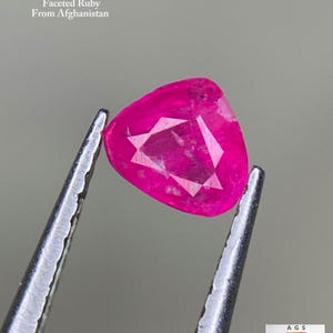 Puede incluir: Una gema de rubí facetada triangular, sujeta con pinzas. La gema es de color rosa vibrante y tiene facetas internas. El texto dice: "0,32 ct Rubí facetado de Afganistán."