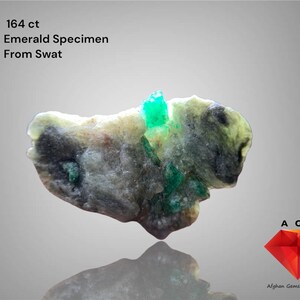 May include: A natural emerald specimen from Swat, Pakistan, with a striking green colour. The stone is a rough-cut formation, displaying a mix of grey, white, and green shades. The image includes the text "164 ct Emerald Specimen From Swat."