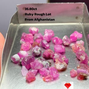 May include: A selection of rough ruby gemstones in a metal tray. The rubies are various shades of pink and red, with some showing white and grey inclusions. Text on the tray reads "35.80ct Ruby Rough Lot From Afghanistan."