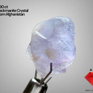 Puede incluir: Un cristal de hackmanita de color púrpura claro de Afganistán, sujeto con pinzas metálicas. La piedra pesa 5,90 ct y tiene un aspecto translúcido. La imagen también incluye el texto "5.90 ct Hackmanite Crystal From Afghanistan" y un logotipo.