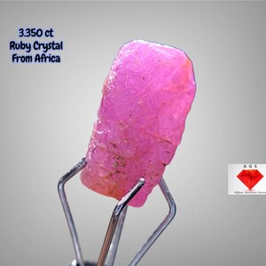 Könnte beinhalten: Ein leuchtend rosa Rubinkristall aus Afrika, gehalten von einem Metallwerkzeug. Der rohe Edelstein hat eine leuchtend rosa Farbe mit einer strukturierten Oberfläche. Der Text auf dem Bild lautet "3.350 ct Ruby Crystal From Africa."