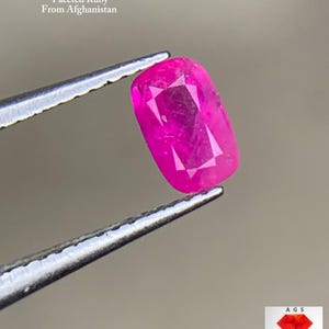 May include: A faceted, rectangular ruby gemstone held by tweezers. The vibrant pink gem, weighing 0.32 ct, is from Afghanistan. The image includes text indicating the gem's origin and weight.