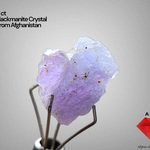 Puede incluir: Cristal de hackmanita de 5 ct de Afganistán, sujeto por un soporte metálico. El cristal tiene una textura rugosa y muestra tonos de lavanda y azul. La imagen incluye el texto "5 ct Hackmanite Crystal From Afghanistan".