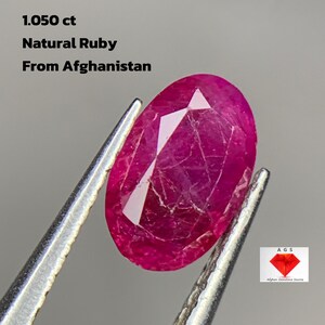 May include: A vibrant, oval-cut natural ruby from Afghanistan, weighing 1.050 ct, held by tweezers. The gemstone is a deep, rich red with visible facets. The image includes text: "1.050 ct Natural Ruby From Afghanistan" and a logo.