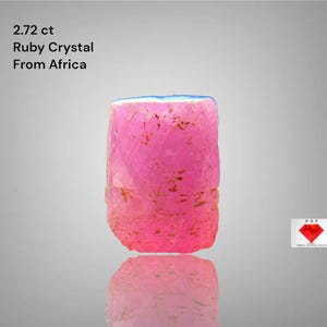Puede incluir: Cristal de rubí rosa vibrante de África, con un peso de 2,72 ct. La gema tiene forma rectangular con una textura ligeramente rugosa y una superficie reflectante. El borde superior tiene un tono azul.