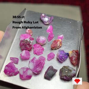 May include: A collection of rough ruby gemstones, ranging in shades of pink, red, and purple, displayed in a metal tray. The text on the image reads "38.55 ct Rough Ruby Lot From Afghanistan."