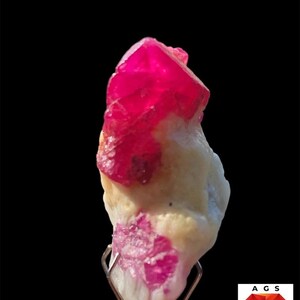 Pode incluir: Um espécime de rubi de 37,2 ct com estruturas cristalinas rosa-vivas incrustadas numa matriz mineral esbranquiçada. A gema bruta é mantida por um suporte metálico, com o texto 37.2 ct Ruby Specimen visível na parte superior da imagem.
