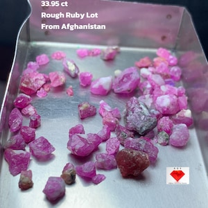 May include: A collection of rough ruby gemstones in a metal tray. The gemstones are various shades of pink and red. Text on the image reads "33.95 ct Rough Ruby Lot From Afghanistan."