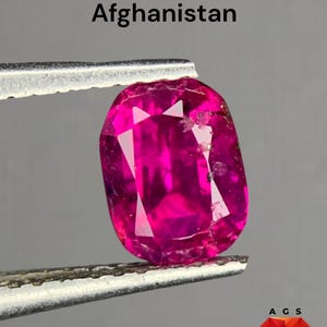 Pode incluir: Um rubi oval vibrante, segurado por pinças, exibe um tom magenta profundo. O texto diz "0,96 carat Ruby from Afghanistan". A gema está sobre um fundo cinza neutro.