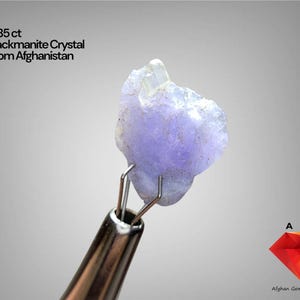 Puede incluir: Un cristal de Hackmanita de color púrpura claro de Afganistán, sostenido por una herramienta de metal. La imagen incluye texto que indica que el cristal pesa 5,85 ct. El logotipo de Afghan Gemstone Source está en la esquina inferior derecha.
