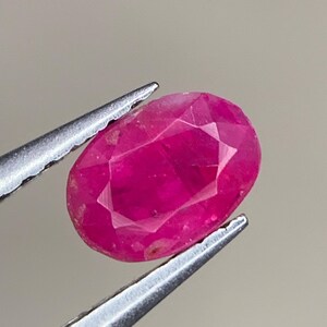 May include: A vibrant pink oval-shaped ruby gemstone with facets, held by tweezers.