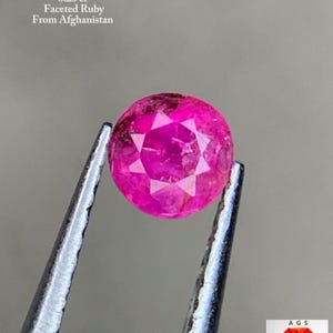 Op de afbeelding: Een gefacetteerde, ronde robijn, vastgehouden door een pincet. De robijn heeft een levendige roze kleur met zichtbare interne facetten. De tekst in de afbeelding luidt: "0,23 ct Gefacetteerde Robijn Uit Afghanistan."