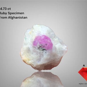 Może przedstawiać: Okaz rubinu o masie 14,73 ct z Afganistanu. Kamień szlachetny ma żywy, różowy środek z rubinu osadzony w białej i brązowej matrycy. Obraz zawiera tekst "Ruby Specimen From Afghanistan".