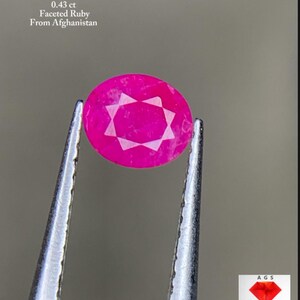 Puede incluir: Un rubí facetado de forma ovalada, sujeto con pinzas. La piedra preciosa es de color rosa vibrante con un patrón en forma de estrella en el centro. El texto de la imagen dice: "0.43 ct Rubí Facetado De Afganistán."