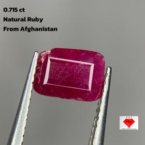 Puede incluir: Un rubí natural de corte rectangular de Afganistán, con un peso de 0,715 ct, se sujeta con unas pinzas. La gema es de color rojo intenso con una superficie pulida. El texto de la imagen dice: 0,715 ct, Rubí natural, De Afganistán.