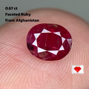 Peut inclure: Une pierre précieuse de rubis facetté d'Afghanistan, pesant 0,67 ct. La gemme est d'un rouge profond avec des facettes blanches, et est tenue entre les doigts. Le texte sur l'image indique : "0,67 ct, Faceted Ruby, From Afghanistan."