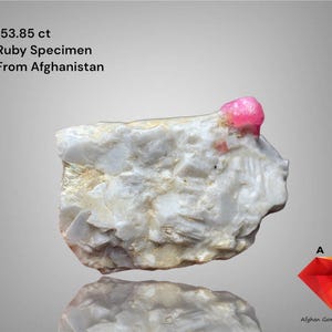 Peut inclure: Spécimen de rubis de 153,85 ct provenant d'Afghanistan. La pierre brute blanche présente un cristal de rubis rose vif. Le texte est clairement visible.