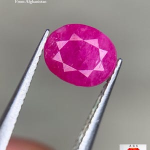 Pode incluir: Uma gema de rubi facetada de cor magenta do Afeganistão, segurada por pinças. A pedra oval tem um padrão de estrela. O texto diz: "1,93 ct Rubi Facetado do Afeganistão."