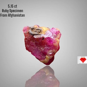 Op de afbeelding: Een ruw, ongeslepen robijn exemplaar uit Afghanistan, 5,15 ct. De edelsteen is een levendig roze-rood met gele en bruine gebieden. De tekst "5.15 ct Ruby Specimen From Afghanistan" is inbegrepen.
