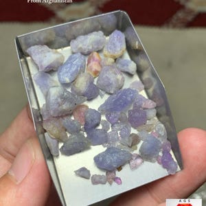 May include: A metal tray holds a collection of rough-cut hackmanite gemstones. The stones, ranging in size, display shades of lavender, purple, and pink. Text at the top reads "58.70 ct Hackmanite Rough Lot From Afghanistan."