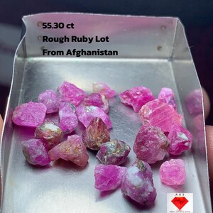 May include: A selection of rough ruby gemstones, weighing 55.30 carats, displayed in a metal tray. The rubies are various shades of pink and red. Text on the tray reads "Rough Ruby Lot From Afghanistan."