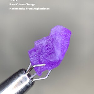 May include: A vibrant purple hackmanite gemstone from Afghanistan, held by a metal tool. The image includes text that reads "1.73 ct Rare Colour Change Hackmanite From Afghanistan."
