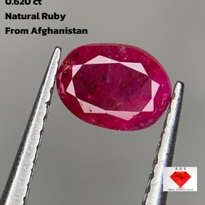Puede incluir: Una piedra preciosa de rubí natural de Afganistán, sujeta con pinzas. La gema roja oscura de forma ovalada tiene una superficie facetada. El texto de la imagen dice: "0.620 ct Natural Ruby From Afghanistan."