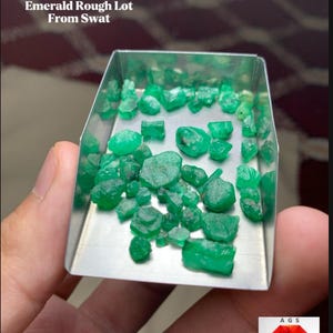 Puede incluir: Un conjunto de esmeraldas en bruto, con un total de 40,35 ct, exhibidas en una pequeña bandeja metálica rectangular. Las piedras preciosas son de color verde vibrante y parecen sin tallar. El texto dice "Emerald Rough Lot From Swat".