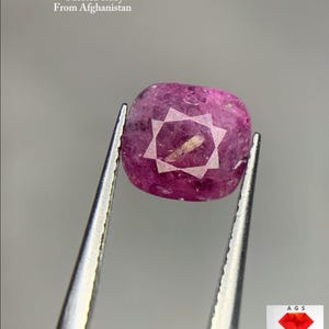 May include: A faceted ruby gemstone, held by tweezers, with the text "2.74 ct Faceted Ruby From Afghanistan". The ruby is a deep pink color with a star-like pattern on its surface. The gemstone is from Afghanistan.