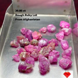 Puede incluir: Un lote de piedras preciosas de rubí en bruto, con un peso de 36,95 ct, exhibido en una bandeja de metal. Los rubíes son de varios tonos de rosa y rojo. El texto en la bandeja dice "Rough Ruby Lot From Afghanistan."