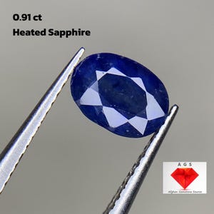 May include: A 0.91 carat heated sapphire gemstone, oval shaped, with a deep blue color. The sapphire is held in tweezers against a white background. The image also includes the text "AGS Afghan Gemstone Source".