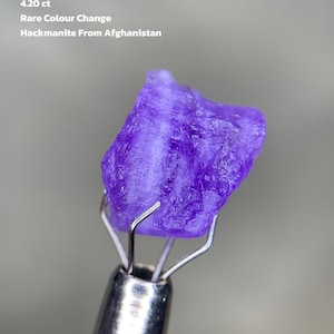 Puede incluir: Una vibrante gema de Hackmanita púrpura de Afganistán, sostenida por una herramienta plateada. El texto dice "4.20 ct Rare Colour Change Hackmanite From Afghanistan". La gema tiene una superficie rugosa y texturizada.