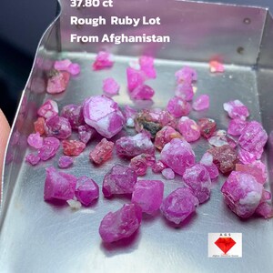 Puede incluir: Una colección de gemas de rubí en bruto en una bandeja de metal. Las gemas son de varios tonos de rosa y rojo. El texto en la bandeja dice "37.80 ct Rough Ruby Lot From Afghanistan."