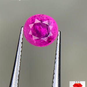 Może przedstawiać: Fasetowany, okrągły, jaskraworóżowy kamień szlachetny rubinu, trzymany przez pęsetę. Kamień ma 0,85 ct i pochodzi z Afganistanu. Kamień ma wzór gwiazdy w środku. Obraz zawiera również logo AGS.