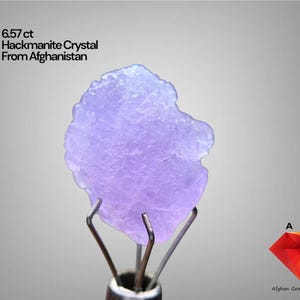May include: A light purple Hackmanite crystal from Afghanistan, held by metal prongs. The crystal is 6.57 ct and has a textured surface. Text on the image reads "6.57 ct Hackmanite Crystal From Afghanistan".
