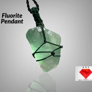 May include: A green fluorite crystal pendant wrapped in dark green cord. The pendant is hanging from a dark green cord. The text "Fluorite Pendant" is in the top left corner of the image. The text "AGS Afghan Gemstone Source" is in the bottom right corner of the image.