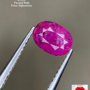 May include: A faceted, oval-shaped ruby, held by tweezers, with a weight of 0.70 ct. The gemstone is a deep pinkish-red color and has a star-like pattern in the center. Text on the image reads "Faceted Ruby From Afghanistan."