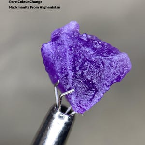 Puede incluir: Una vibrante gema de Hackmanita púrpura de Afganistán, con un peso de 6,21 ct. La rara piedra que cambia de color está sujeta por una herramienta de metal plateado. El texto en la imagen dice "Rare Colour Change" y "Hackmanite From Afghanistan."