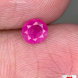 Könnte beinhalten: Ein facettierter, runder Rubin-Edelstein, 0,45 ct, aus Afghanistan. Der Edelstein hat eine leuchtende rosa-rote Farbe mit einem sternförmigen Facettenmuster. Der Text "0.45 ct Faceted Ruby From Afghanistan" ist ebenfalls zu sehen.