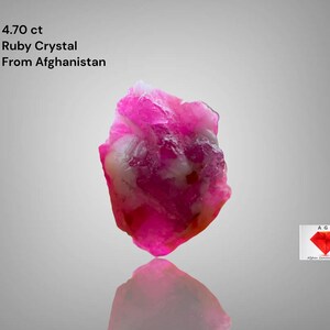Può includere: Un vivace cristallo di rubino rosa dall'Afghanistan, del peso di 4,70 ct. La gemma ha una forma ruvida e irregolare con aree bianche e una superficie riflettente. Il testo "4.70 ct Ruby Crystal From Afghanistan" è incluso.