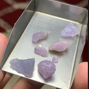 May include: A selection of rough hackmanite gemstones displayed in a metal tray. The stones vary in colour from pale pink to lavender and purple. The image includes text: "22.20 ct Hackmanite Rough Lot From Afghanistan."