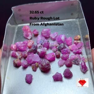 Puede incluir: Una colección de gemas de rubí en bruto en una bandeja de metal. Las gemas son de varios tonos de rosa y rojo. El texto en la bandeja dice "32.65 ct Ruby Rough Lot From Afghanistan". Un logotipo de Afghan Gemstone Source está en la esquina inferior derecha.